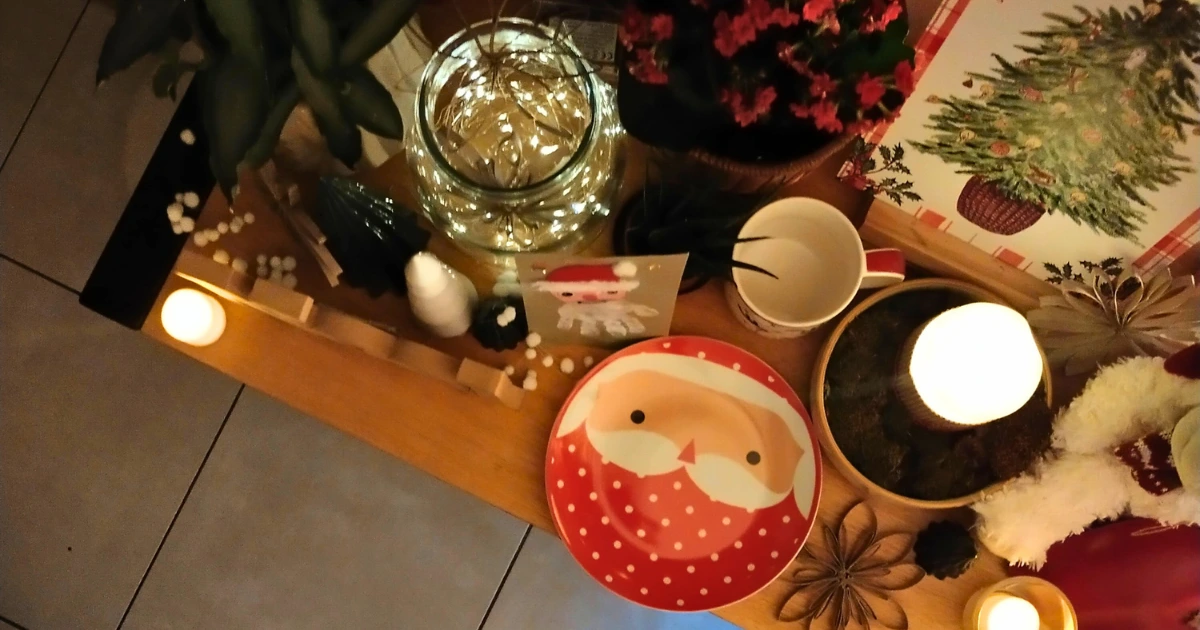 Décor de Noël chaleureux sur une table : assiette et carte Père Noël, mug, bougie allumée, plantes, guirlande lumineuse et petites décorations festives.
